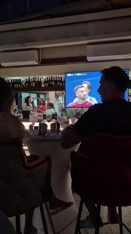 A man and a woman are seated at a bar watching a soccer game on a large television screen. The bar has a shelf stocked with bottles behind the counter, where several people are standing. The room is dimly lit and appears to be a restaurant or bar.