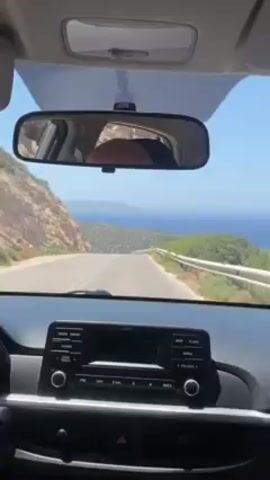 The video shows a view from inside a car driving on a coastal road. The road curves along a hillside covered in green vegetation. Beyond the road, the blue ocean stretches to the horizon, where islands can be seen. The car's dashboard is visible, including the radio and gear stick. A person's head is visible in the rearview mirror. Another car appears on the road ahead.