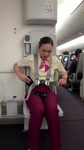 A flight attendant, dressed in a cream-colored uniform shirt with a purple and orange scarf and purple pants, sits on an airplane jump seat, demonstrating how to fasten the safety harness. She adjusts the straps, connecting them and tightening them around her torso. The jump seat is positioned against a white wall with a patterned design. To the left of the jump seat is a trash receptacle. In the background, rows of airplane seats are visible, some occupied by passengers. The flight attendant's hair is pulled back into a bun.