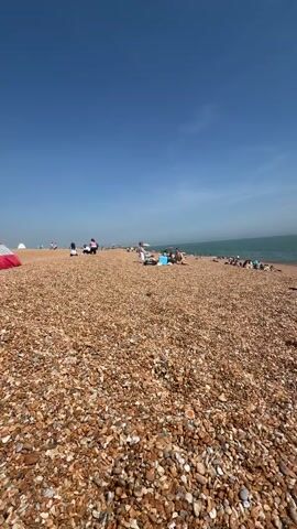 The video depicts a pebble beach on a sunny day, with numerous people scattered across the shoreline. Some individuals are lying down on towels or blankets, possibly sunbathing. Others are sitting in groups, chatting or relaxing. A few people are standing or walking along the beach. In the background, the calm sea stretches out to the horizon under a clear blue sky. The beach is entirely covered in pebbles, ranging in color from light tan to darker brown.