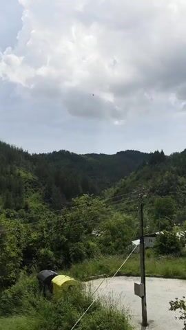 The video presents a panoramic view of a lush, mountainous landscape in Rudozem, Bulgaria, under an overcast sky. The shot slowly pans from a slightly left-of-center position to the right. In the foreground, a utility pole with multiple wires stands prominently, casting a shadow. Below it, a paved area, resembling a road or parking lot, is visible, bordered by overgrown green grass and bushes. A small, partially visible yellow structure, possibly a tent or canopy, is situated to the far left.

The mid-ground and background are dominated by rolling hills and mountains densely covered in vibrant green trees, indicating a heavily forested area. Scattered among the trees and along the base of the hills, several small houses or buildings can be seen, suggesting a semi-rural setting. The sky is largely covered by dark, heavy clouds, implying a potentially overcast or rainy day, though no rain is visible.

Throughout the entire video, a loud, continuous siren sound is heard, characterized by its fluctuating pitch, typical of an air raid or emergency warning siren. The sound provides a stark contrast to the otherwise tranquil visual scene, evoking a sense of urgency or alert. There are no visible people or discernible text in the video. The time of day appears to be daytime.
