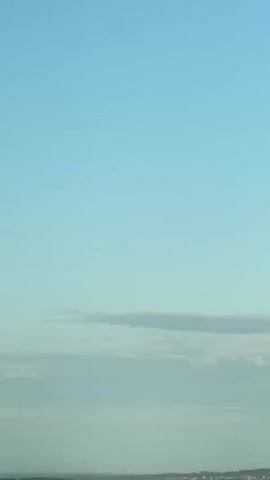 A vertical, wide shot captures a pale blue sky with a few thin, elongated clouds. Below the sky, a hazy horizon is visible, with a dense line of green trees and some structures that appear to be buildings and possibly industrial facilities. The foreground is dominated by the top of a modern building with white and teal-colored sections. The camera is static.