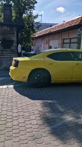 A yellow Dodge Charger is parked on a cobblestone street in front of a building with a banner that reads "RYBY WĘDZONE PRZETWORY RYBNE" (Smoked fish fish products). A man on a bicycle wearing a helmet and backpack rides past the car. The weather is sunny with some clouds.