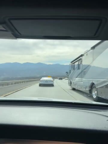 The video shows a first-person view from inside a vehicle traveling on a highway. The road is paved with multiple lanes, separated by solid white lines. In front, there is a white sedan with a California license plate and a yellow sign on its roof. To the right, a large RV is visible. Beyond the highway, there are mountains in the background under a cloudy sky. A yellow sign with black text is seen on the side of the road, reading "RUNAWAY TRUCK RAMP 2 MILES." The vehicle appears to be in traffic, moving at a moderate pace. The overall environment suggests a mountainous or hilly region.