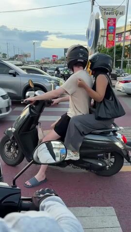 Two people, a male driver and a female passenger, are riding a black scooter. They are both wearing helmets and appear to be stopped at a crosswalk, waiting for traffic to pass. The male is wearing a tan t-shirt and black shorts, while the female is wearing a black tank top and gray pants. She has a black purse slung over her shoulder. The scooter is a modern, sleek design.

The scene is an urban street in Chiang Mai City Municipality, Thailand. Cars and other scooters are visible in the background, suggesting a busy intersection. A pedestrian crossing signal is visible, indicating they are at a marked crosswalk. The sky is cloudy, suggesting it might be late afternoon or early evening. The overall atmosphere appears to be one of routine travel within a bustling city.

Notable details include the "Central Festival" sign visible in the background, a large shopping mall complex, which helps to pinpoint the location. The scooter has the brand name "Vino" visible on the side. The subjects' posture and the way they are positioned on the scooter suggest comfort and familiarity with riding together. The reflection in the scooter's side mirror shows a person on another scooter from a different perspective. The colors are muted, with the sky being a pale blue and gray, and the road a muted red and white.