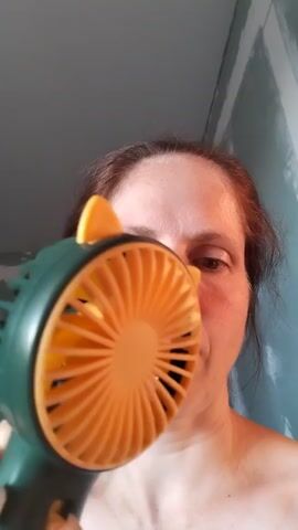 A woman holds a small, green and orange fan to her face and the fan appears to be on as the blades are spinning. The woman's eyes are closed. The fan is plugged in to a white cord. The background appears to be a room with light green walls.