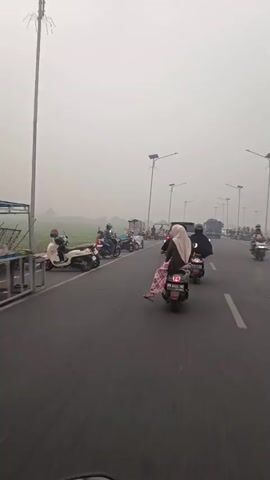 The video shows a road scene with several motorcycles and trucks. In the foreground, a person is riding a scooter, wearing a hijab and a patterned skirt. Another person is riding behind them on the same scooter. To the left, other motorcycles are parked, and several people are standing around them, some near food stalls. In the background, a truck is driving on the road, and the sky is hazy or overcast. The overall environment appears to be a rural or semi-rural area with agricultural fields visible on the left side of the road. The video has a slightly shaky camera movement, suggesting it was filmed from a moving vehicle.