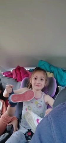 A young girl is sitting in a car seat in the back of a car, smiling and playing with her shoes. She is wearing a grey tank top with cartoon animals and grey pants. The car seat is purple and grey. Behind her are jackets and other items stacked on the back seat. Another child is partially visible to her left. Someone wearing a blue shirt is in the foreground, likely the person filming.  The girl holds up each shoe, showing the soles, and then she puts one back on.  There is music playing in the car.