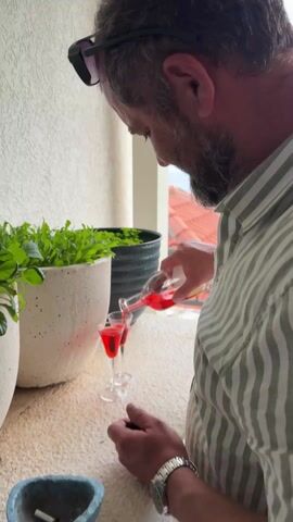 A man with a beard wearing a striped shirt is pouring a red liquid from a glass bottle into two shot glasses on a white ledge. He then smiles at the camera and holds up the bottle in his fist. He is standing on a balcony with plants in pots on the ledge. In the background, there is a view of red rooftops and the ocean. The man is wearing sunglasses on his head and a silver watch on his wrist.