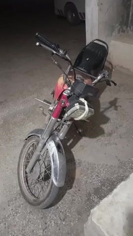 A single, static, eye-level shot shows a parked motorcycle, primarily red and silver, facing the camera. The motorcycle's headlight appears to be taped together. The tires appear worn, and the overall condition is somewhat weathered.  The ground is a speckled gray. In the background, the corner of a white vehicle is visible. The scene seems to be outdoors at night, with a concrete structure bordering the right side of the frame.