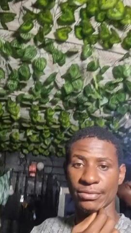 A close-up video shows a young Black man looking into the camera. He has short, dark hair and is wearing a gray striped shirt and a black wireless earbud in his right ear. He briefly touches his chin with his right hand, makes a kissing face, and then smiles slightly. The background features a dense arrangement of artificial green leaves covering the wall and ceiling. In the blurry background, other furniture, possibly a TV, a mirror, and a bottle of pink liquid are visible.