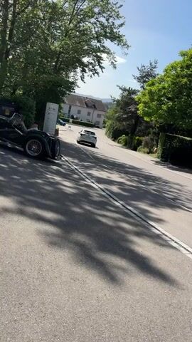The video shows a vintage dark green convertible car driving down a paved road. The car has two occupants. The driver is on the left side of the vehicle and appears to be a person wearing a light-colored shirt. The passenger is on the right side and is also wearing a light-colored shirt. The car's wheels have red accents. The road is bordered by green trees and hedges. In the background, there is a white building with a red roof and another white car. The sky is blue with some white clouds, suggesting a sunny day. The camera is positioned at a low angle, looking up at the car as it drives away. The video is approximately 5 seconds long.