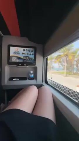 A person sits next to a large window in what appears to be a modern, luxurious bus or private vehicle, looking out at a beach scene. The window shows a palm tree-lined road with a beach and ocean in the background under a bright, sunny sky. The person, dressed in a black skirt, sits with their knees bent. Beside them is a panel with a small screen displaying what looks like a menu of food and beverages. There’s also a small bottle of water and a phone resting on a shelf. The interior has dark gray walls with a red accent stripe on the ceiling. The overall setting is comfortable and upscale, suggesting a leisurely journey with a scenic view.