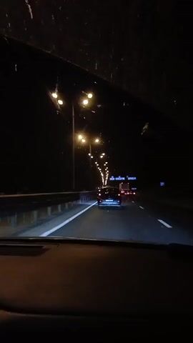 The video is taken from inside a vehicle, looking out through the windshield at a highway at night. Streetlights illuminate the road, and other vehicles can be seen driving ahead. The windshield has some marks and smudges on it. The dashboard of the car is visible in the foreground.