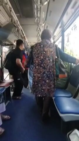 The video shows the interior of a bus with several passengers. The camera is moving and the scene is slightly blurry. In the foreground, a person wearing a floral shirt and a blue cap is standing and holding onto a handrail. To the left, another person is partially visible, wearing a patterned skirt and a light-colored top, also holding a handrail. Further back, other passengers are visible, some sitting and some standing. One person in the background is wearing a floral dress. A woman in a green headscarf is seated by the window. The bus has blue seats and metal handrails. The floor of the bus is blue. The overall impression is of a crowded public transportation vehicle.