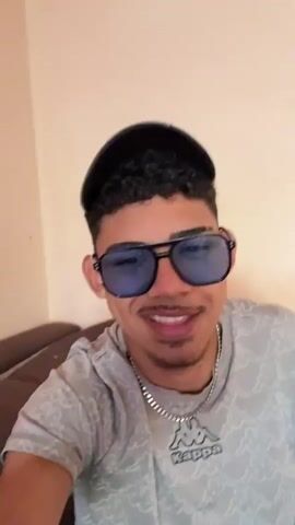 This is a selfie-style video clip of a young man wearing sunglasses, a cap, and a t-shirt. He is smiling and appears to be indoors, possibly in a living room, as indicated by a wall socket visible in the background. The lighting suggests daytime. The man is wearing a silver chain necklace with a Kappa logo. The background wall is a light peach color, with a hint of pink and yellow abstract art on the right side. The man's skin tone is light brown, and he has curly dark hair. His facial expression is happy and relaxed. The clip is short, with no discernable dialogue or significant events taking place beyond the man's pose for the camera.