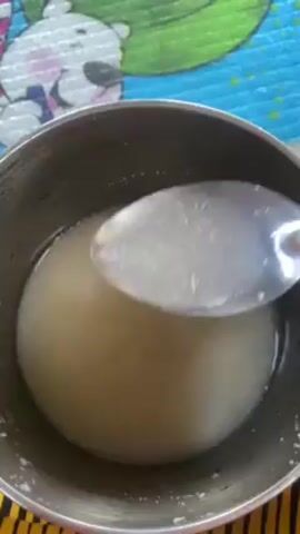 A top-down shot shows a stainless steel pot containing a cloudy, opaque liquid. A black spoon is used to stir the contents. The pot is sitting on a surface decorated with a pattern of a cartoon polar bear and other colorful shapes. The light reflects off the surface of the liquid, creating a bright, diffused glow within the pot.