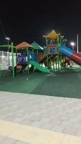 The video shows a children's playground at night. A brightly colored plastic playground structure with multiple slides and platforms is the central focus. The ground is covered in green artificial turf, with a patterned paved area in the foreground. The sky is dark, and artificial lights illuminate the playground. In the background, a few blurred figures are visible, suggesting the presence of people. The camera pans slowly across the scene.