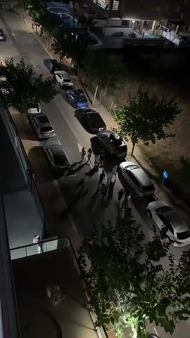 This is a night-time aerial view of a residential street in Les Amplaries, Spain, lined with parked cars and trees. A group of people, estimated to be around eight to ten individuals, are gathered in the middle of the street, appearing to be interacting or conversing. The scene is illuminated by streetlights and the ambient light from nearby buildings, casting long shadows on the asphalt. The time of day is clearly night, as indicated by the darkness and artificial lighting. The weather appears to be clear, with no visible rain or fog. The buildings visible in the background suggest a typical European urban or suburban environment. The overall mood is relaxed, with the group appearing to be engaged in a casual social gathering. There is no visible text in the media file.