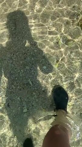 The video shows a shadow of a person walking through shallow, clear water. The person's feet are visible as they step into the water, and the shadow reflects the person's silhouette as they hold something up to their head. The water bottom is visible, covered with small stones and pebbles. The light shimmers on the water's surface, creating a dappled effect on the seabed and the shadow.