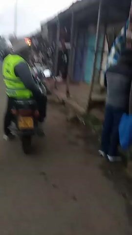 A person in a gray jacket and dark pants walks on a street alongside parked motorcycles and small businesses. The person has a large white bag slung over their shoulder. A person in a yellow safety vest and black pants is seated on one of the motorcycles. The street appears busy.
