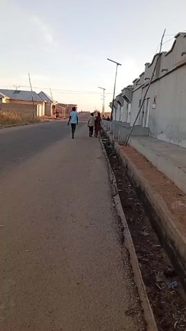 The video captures a street scene in what appears to be a residential area. A paved road stretches into the distance, flanked on one side by a row of identical, modern-looking houses with light gray walls. These houses have solar-powered streetlights in front of them. A concrete drainage ditch runs along the side of the road, filled with dirt and some litter.

Several people are walking down the road away from the camera. A man in a blue shirt walks farthest ahead, while a group of two or three individuals follow behind him. In the distance, another person can be seen walking.

The sky is light blue, suggesting it is either early morning or late afternoon. The lighting is fairly even, casting soft shadows. The overall impression is of a quiet, somewhat sparse, residential neighborhood.