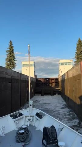 A boat is moving towards a large metal gate at the end of a canal lock. The water in the lock is turbulent with white foam, indicating water is flowing. The lock walls are made of concrete with some moss or algae growth. Two white buildings with blue accents are visible on either side of the lock. The sky is blue with some white clouds. Two evergreen trees are visible on the left and right sides of the lock entrance. The bow of the boat is in the foreground. The boat has white paint and some ropes are visible on its deck.