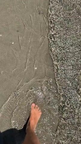 The video shows a first-person view of a person walking barefoot along the edge of the ocean. The camera is angled downwards, focusing on the person's legs and feet as they step into and out of the shallow, clear water. The sand is visible along the left side of the frame, wet and darker where the water has receded. The water itself is shallow, with small waves gently washing over the sand. Sunlight sparkles on the surface of the water. The person is moving from left to right across the frame and slightly away from the camera. Their skin tone appears to be tanned.