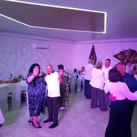 The video shows a dimly lit indoor event space, likely a banquet hall, decorated with white tablecloths on tables, chairs, and a textured wall. A group of people are dancing and socializing. In the foreground, a woman in a dark patterned dress and a man in a white shirt and dark pants are dancing. A woman and a man are dancing to their right, with the woman wearing a white top and black pants. Further back, several other couples and groups of people are visible, some standing and talking, others dancing. The lighting is a mix of white and colored ambient light, with a prominent rectangular light fixture on the ceiling. The overall atmosphere appears to be festive, possibly a wedding reception or a similar celebration.
