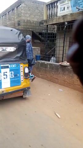 The video begins with a close-up view of the interior of a yellow auto-rickshaw. A person, wearing a maroon top and green pants, is seated inside, their body angled away from the camera. The auto-rickshaw has a black canopy overhead. The camera then pans to the right, revealing a street scene. On the right side of the street, there are several small shops or stalls with open fronts, displaying various goods. A low concrete wall separates the shops from the street. The ground is unpaved and appears to be dirt or sand, with some scattered debris. In the distance, more buildings and structures are visible, suggesting a bustling urban or semi-urban environment. People are seen walking on the street and near the shops. The overall impression is of a lively marketplace or street scene.