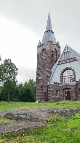 The video shows a stone church with a tall spire. The church is made of large, rough-hewn stones, primarily in shades of reddish-brown and grey. The spire is grey and pointed, with several tiers. To the right of the spire is the main body of the church, which has a gabled roof covered in grey metal panels. The facade features a large arched window above the entrance. There are several smaller windows on the lower level. The church is surrounded by green grass and some trees. The sky is overcast and grey. The video pans slightly to the right, revealing more of the church's side and the surrounding greenery. There are no people visible in the video.