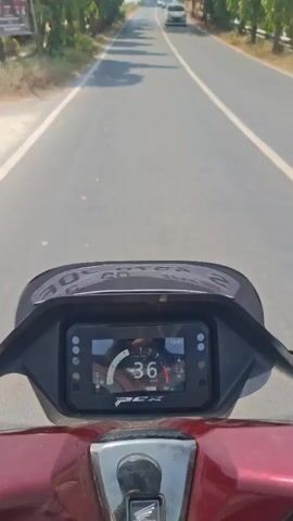 The video begins with a close-up of a motorcycle's digital dashboard, showing a speed of 37. The camera then pans up and out, revealing a view from the motorcycle as it travels down a paved road lined with trees and sparse vegetation. The road appears to be in a rural or suburban setting, with a few cars visible in the distance. The sky is bright and sunny. The motorcycle is moving at a moderate speed. There are no other people clearly visible in the frame other than the rider, whose hands are on the handlebars. The video captures the act of riding a motorcycle on a clear day, with a focus on the road ahead and the motorcycle's instrumentation.
