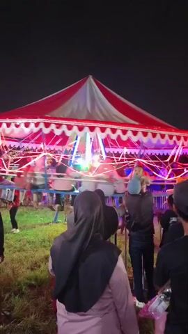 The video shows a brightly lit carousel at night. The carousel has a red and white striped canopy and is decorated with flashing lights. Several people are visible on and around the carousel, including children and adults. Some people are riding the carousel, while others are standing and watching. The ground around the carousel appears to be uneven and covered in grass. The overall atmosphere is festive and lively.