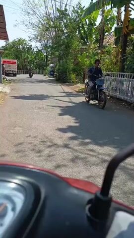 A first-person perspective video shows a person riding a red scooter down a paved road. Trees and houses line both sides of the road. There are other people on scooters and a white truck in the distance. The sun is shining, casting shadows on the road.