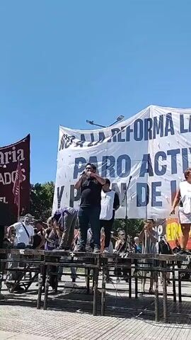 The video captures a protest against labor reform. A group of people stand on a makeshift stage constructed from tables. A man with a microphone, dressed in a black shirt and blue jeans, speaks passionately. A woman in a white top and patterned shorts stands to his right. Behind them, a large banner reads "NO A LA REFORMA LABORAL" (No to labor reform) and "PARO ACTIVO Y PLAN DE LUCHA" (Active strike and plan of struggle). The sky is clear and blue. Other protesters and supporters are present around the stage. The overall tone of the video suggests a political demonstration or rally.