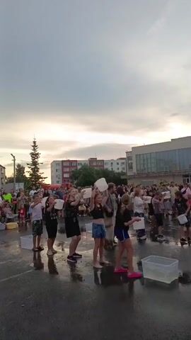 The video shows an outdoor event in Криводановка, Russia. A group of people, mostly children and young adults, are participating in what appears to be an ice bucket challenge or a similar water-based activity. They are standing on a wet asphalt surface, with several clear plastic tubs of water placed in front of them. Each participant holds a white bucket or container above their head, suggesting they are about to pour water over themselves or have just done so.

In the background, a crowd of spectators has gathered, observing the event. The setting includes buildings, trees, and a cloudy sky, indicating it is likely late afternoon or early evening, with overcast weather. The atmosphere seems to be one of fun and participation, with some individuals smiling and engaged in the activity. The ground is visibly wet, reflecting the sky and the participants, reinforcing the water theme of the event.

There is no visible text in the video. The focus is on the action of the participants and the community gathering.