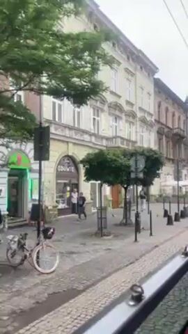 A tram ride showing buildings on a street. The buildings are several stories tall with many windows. The facades have elaborate trim in places. There are trees with rounded tops in front of the buildings, along with street signs and bollards. People are walking on the sidewalk. There are bicycles parked. A computer store and cafe are visible in front of the buildings. The sky is overcast.