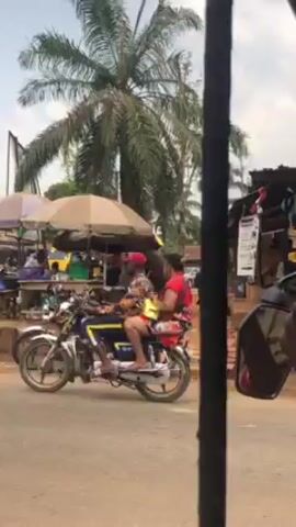 The video depicts a street scene in what appears to be a tropical or subtropical region.  A tall, leaning palm tree dominates the background. In the foreground, two motorcycles are parked near a roadside stall shaded by a large umbrella.  A person is kneeling and working on one of the motorcycles. Another person in a patterned shirt walks past the camera, partially obscuring the view.  A small yellow auto-rickshaw is visible behind the stall. The ground appears to be reddish dirt.