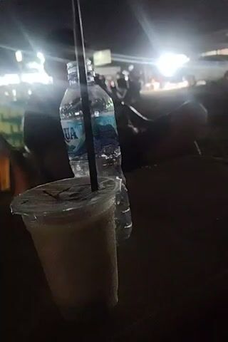 The video shows a night scene at what appears to be an outdoor cafe or gathering space. There's a drink with a straw in a plastic cup and a bottle of Aqua water on the table in the foreground. In the background, there are people sitting in plastic chairs at tables, some under green umbrellas or awnings. Lights are visible overhead, creating a bright, somewhat blurry effect. The overall impression is of a casual, outdoor gathering place at night.