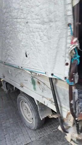A small truck parked on a paved area is seen in the video. The truck's bed has a weathered appearance and is partially covered with a canvas tarp. Several people are seen climbing into the back of the truck. The people are wearing casual clothing, including pants, shirts, and sweaters. The setting appears to be an outdoor area with buildings in the background.