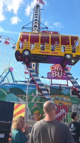 A yellow bus-shaped amusement park ride, labeled "Crazy Bus," is in motion against a bright blue sky with scattered clouds. The ride is suspended from a tall, dark-colored support structure and appears to be spinning or swinging. Several children are visible inside the bus. In the foreground, three adults are standing, with two looking towards the ride and one looking away from the camera. Behind the ride, a Ferris wheel can be seen, along with other amusement park structures and banners. The overall scene depicts a lively atmosphere at a fair or carnival.