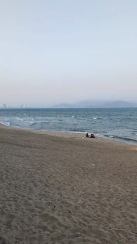 The video shows a beach scene with two people sitting close together on the sand near the water's edge, and other people swimming in the sea. In the distance, there are visible mountains and a city skyline obscured by what appears to be haze or smog. The sky is a pale, muted color, suggesting it might be early morning or late afternoon. Gentle waves are rolling onto the shore.