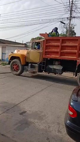 Here's a description of the video:

The video shows an older, rusty yellow and orange dump truck making a slow left turn on a paved street. The truck has a large red dump bed with ribbed sides. A person wearing a dark hooded garment sits atop the load bed, facing forward and appearing to hold something to their face, possibly a phone. The truck's tires are large and dirty, with red wheel hubs.

The street has visible cracks and imperfections. To the right of the frame, a portion of a dark-colored car is visible. Power lines are strung overhead, and a few buildings can be seen in the background. The sky is overcast.