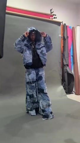 The video shows a fashion shoot in a studio setting. A young man wearing a unique outfit made of patchwork denim—a hooded jacket and wide-legged pants—poses in front of a gray backdrop. The studio has lighting equipment including softboxes on tripods. In a different part of the studio, visible in a mirror, several individuals are preparing. One is adjusting his hair and taking a drink. He is wearing a denim jacket, striped pants, and a studded belt. Others are also visible in the reflection, though their details are less clear. The overall impression is a behind-the-scenes look at a fashion photoshoot.