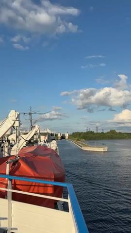 The video captures a view from the deck of a boat as it approaches a lock system. The water is calm, reflecting the blue sky and scattered white clouds. The boat is moving forward, indicated by the wake behind it. On the left side of the frame, part of the boat's deck is visible, including a red-covered life raft. Further ahead, the lock gates are prominent. These are tall, white structures with the number "2" visible on them. The lock is situated within a channel that is bordered by green grassy banks and some trees in the distance. The overall environment suggests a waterway with managed navigation.