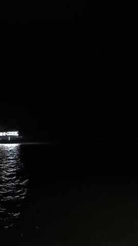 The video shows a large, illuminated ferry or boat moving across a dark body of water at night. The boat's lights are reflected in the water, creating shimmering patterns. The surrounding environment is very dark, with minimal visible details beyond the boat and the water. Some indistinct voices can be heard in the background.