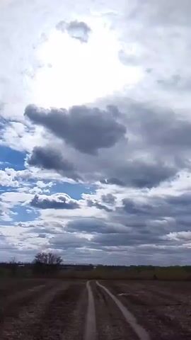The video shows a low angle view of a field with a dirt road leading towards the horizon. The sky is filled with a mix of dark and light clouds, with the sun shining brightly through a gap in the clouds. The wind is audibly strong. A few small trees and bushes are visible along the horizon.