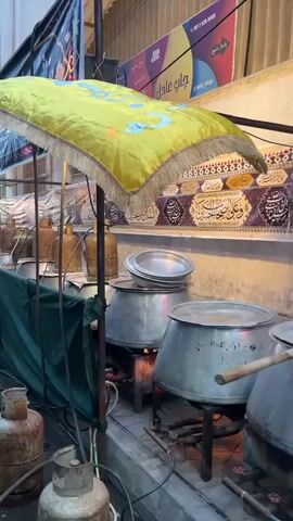 A low-angle medium shot pans to the right across a street food stall at dusk. The stall is set up along a wall adorned with decorative banners and calligraphy. In the foreground, a propane tank with a green hose is visible to the left. Behind it, a green tarp drapes down. Above the tarp, a yellow canopy with gold trim is stretched over the stall. Several large metal cooking pots are arranged on burners in the middle ground. Some pots are covered with lids, while others are open, revealing cooking contents. Gas cylinders, varying in color from gold to brown, are placed on top of the lids of some of the pots, likely for cooking fuel. In the background, the wall is decorated with horizontal bands of Islamic calligraphy and patterned designs. The lighting suggests an evening setting, with artificial lights illuminating the scene.