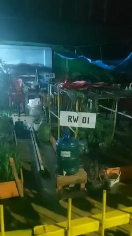 The video shows an outdoor area at night. There are several stands set up, possibly for a market or stalls. Signs with "RW 01" and "RW 03" are visible. In the foreground, there are some yellow barriers. A large green water jug is placed on a wooden stand in front of the "RW 01" sign. Various plants and greenery are present around the stands. The lighting is dim, with some bright lights illuminating the stalls, creating a somewhat blurry and hazy effect. The overall impression is of a temporary outdoor setup, possibly a nighttime market or vendor area.
