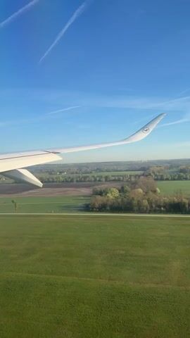 The video shows a view from inside an airplane as it is landing. The wing of the Lufthansa plane is visible on the left side of the frame. Below, a green grassy field stretches out, bordered by trees and what appears to be agricultural land. The sky above is a clear blue with faint contrails. A small, orange structure sits on the edge of the field. The camera is stationary, capturing the steady approach to the ground.